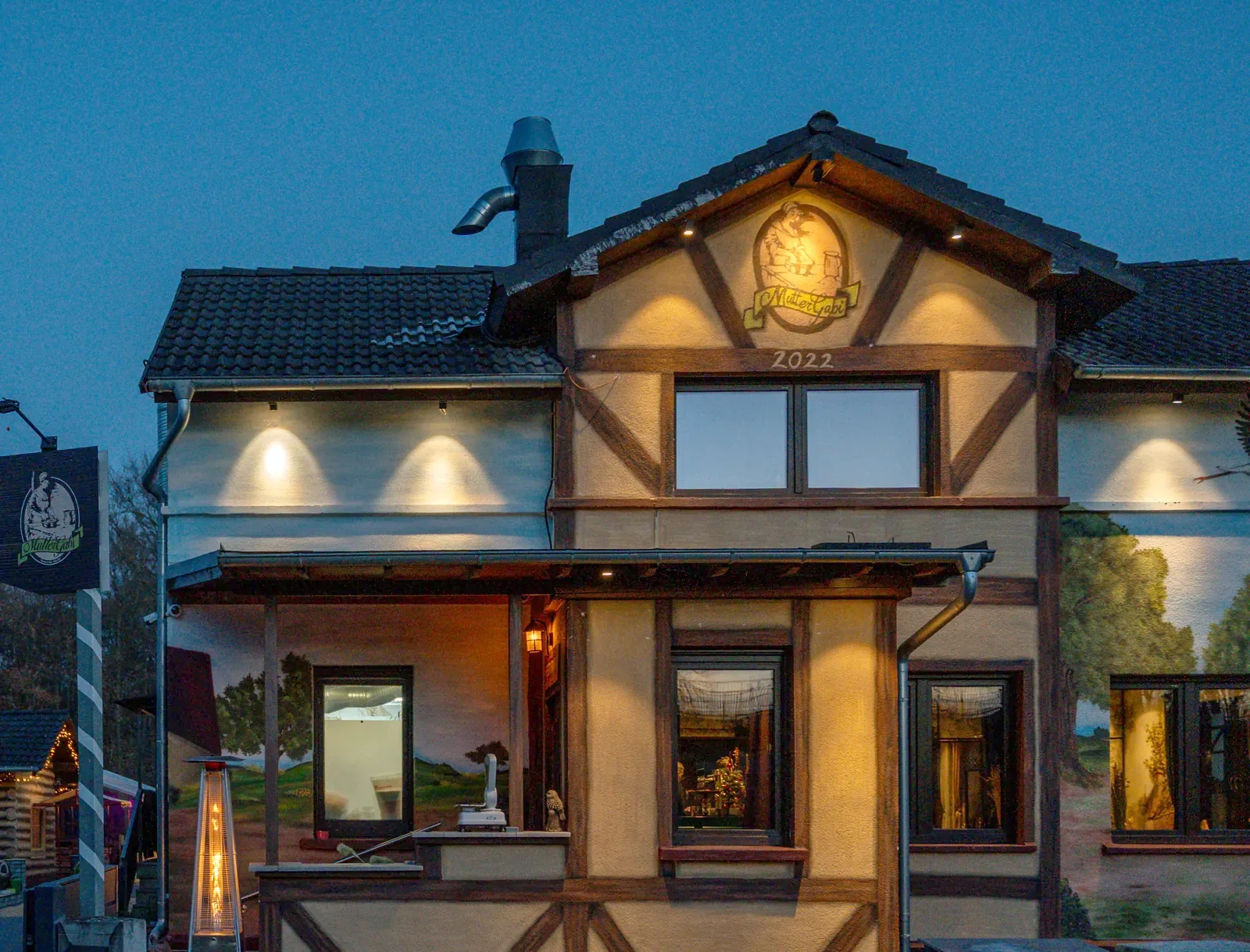 Das Fachwerkhaus von Mutter Gabi mit beleuchtetem Logo am Giebel zur blauen Stunde