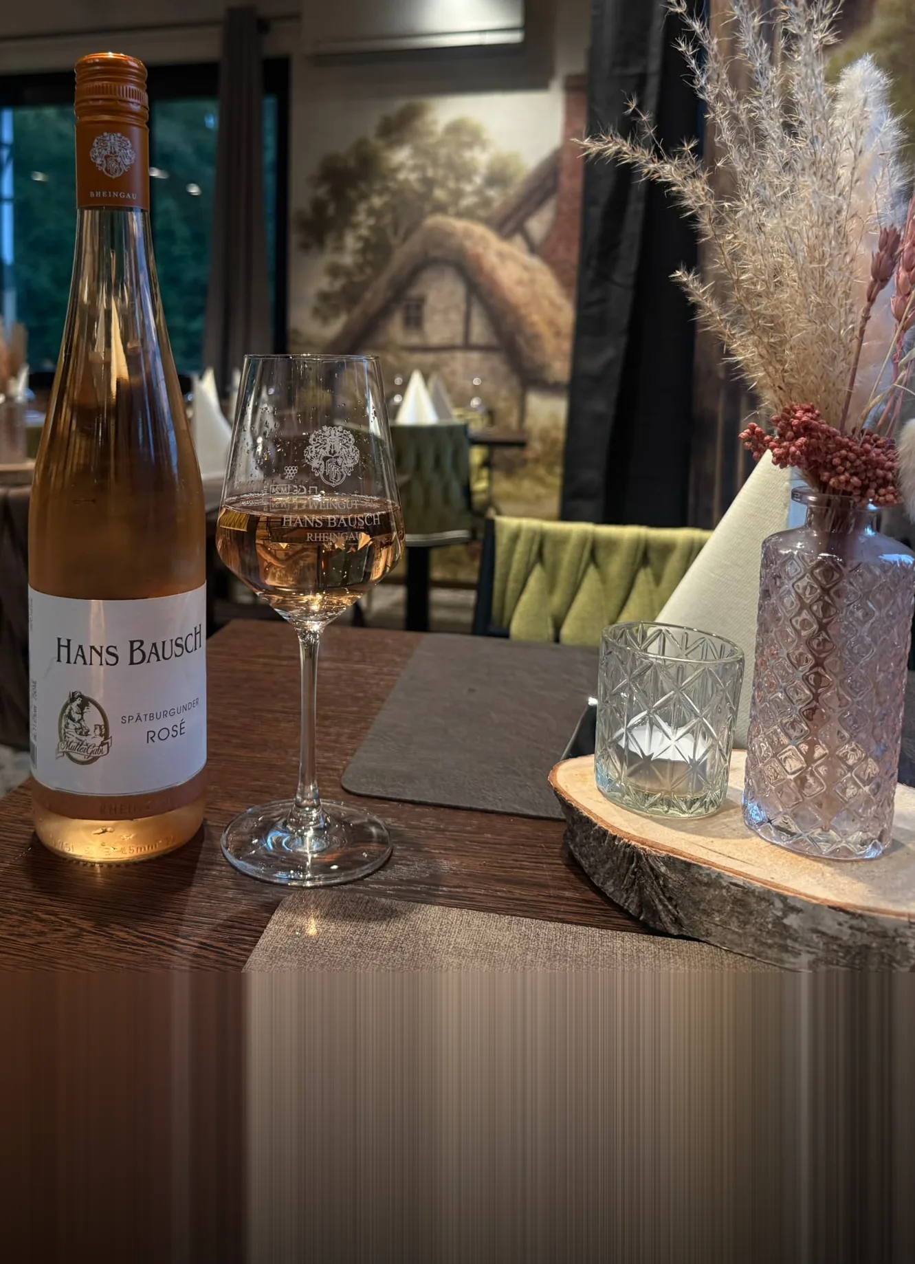Holztisch im Restaurant mit Trockenblumen, Windlicht und einem Glas Rosé vor dem Wandbild