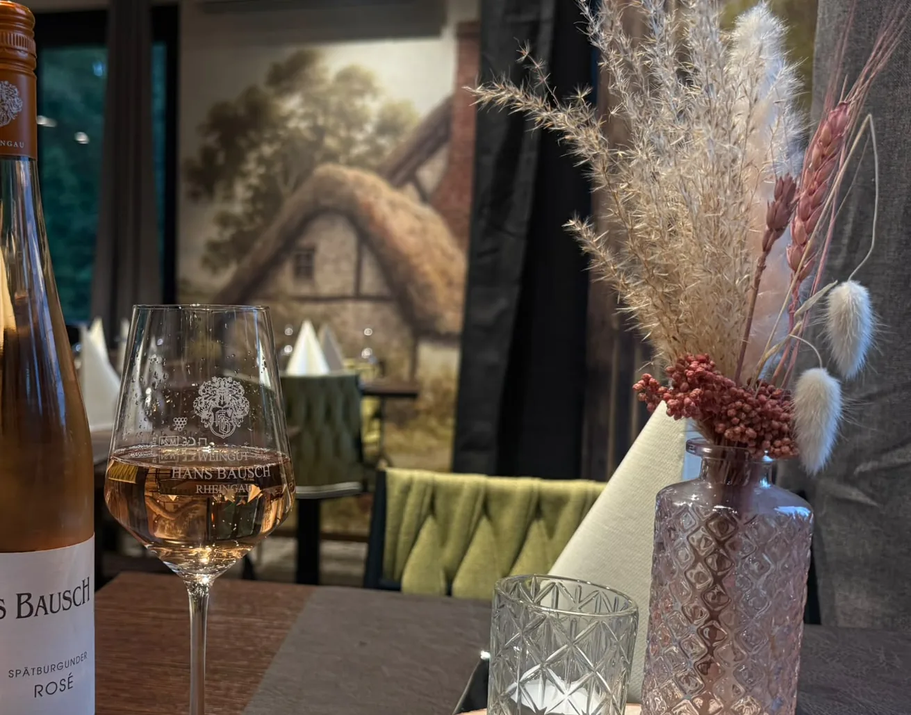 Holztisch im Restaurant mit Trockenblumen, Windlicht und einem Glas Rosé vor dem Wandbild